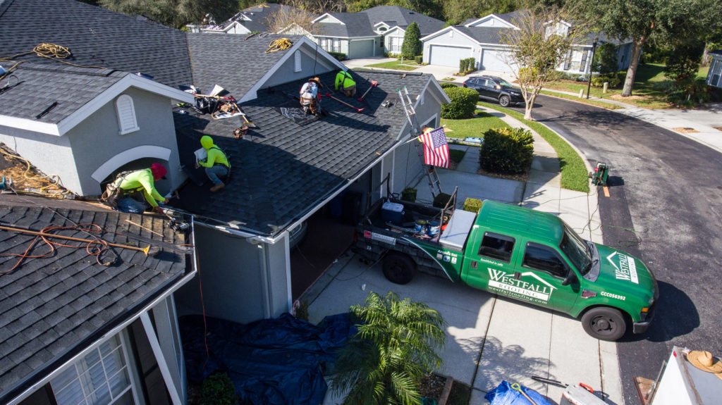 Roofing contractor crew working on a new roof in a residential area, with a Westfall Roofing truck in the driveway.